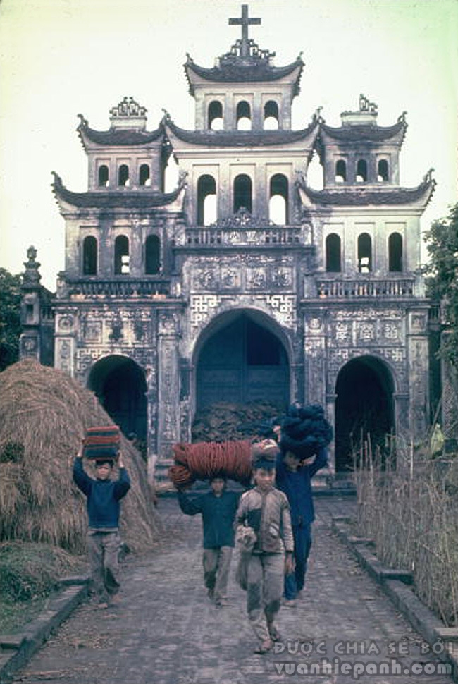 1967. Phía trước một nhà thờ ở Phát Diệm, người lao động khiêng sợi gai dầu dùng để dệt thảm và chiếu.